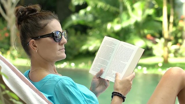 Beautiful woman reading book while sitting on sunbed and smiling to the camera, steadycam shot
