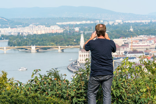 A man standing in green bushes up high at a viewpoint to take a panoramic picture Budapest below.