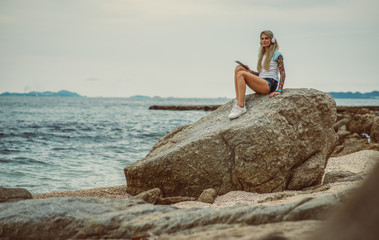 Fototapeta na wymiar Beautiful blonde woman sitting on a rock by the sea and listening to music on stylish headphones on the tablet. hands with a tattoo. The style of summer holiday. Modern girl