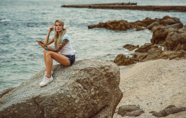 Beautiful blonde woman sitting on a rock by the sea and listening to music on stylish headphones on the tablet. hands with a tattoo. The style of summer holiday. Modern girl
