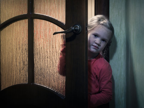 A Child Peeps Standing Behind A Door In A Dark Room