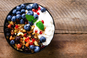 Tasty homemade granola, yogurt, fresh organic berries, pomegranate, mint on rustic wooden background with copy space, top view.