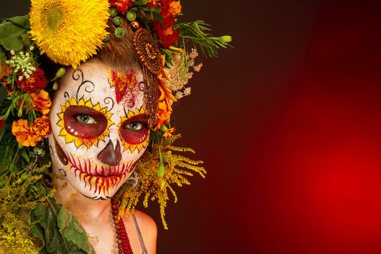 Женщина с разрисованным лицом к мексиканскому Дню Мертвых с цветами на красном фоне.