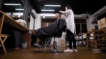 Side view of female young barber standing and doing hairstyling with comb and scissors at the barbershop. Handsome man is sitting on the chair and looking at the mirror opposite.