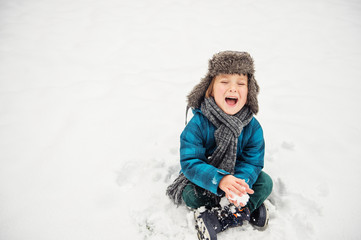 Cute little boy playing in winter park. Kid having fun outdoors, sitting on the snow, wearing warm...