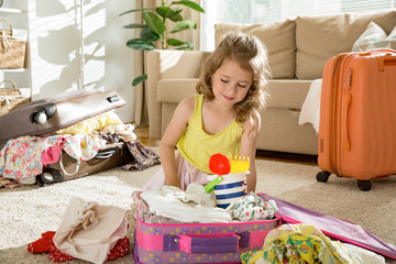 Happy little girl sitting in living room at home, carefully packing colorful summer clothes  and...
