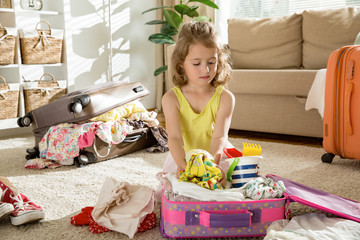 Happy little girl sitting in living room at home, carefully packing colorful summer clothes  and toys into luggage suitcase for a new journey. Child Traveling