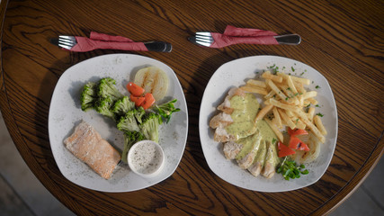 Top view of plates with food from the restaurant, which stand on a wooden table. On the dishes lie a fish with vegetables and chicken with potatoes