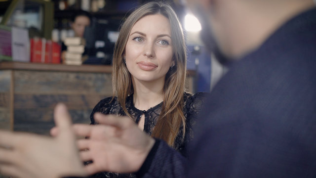 Close up of beautiful woman with long fair hair spending free time in the restaurant. Adorable lady with natural make-up in black lace dress speaking with male hands gesticulating in the foreground.