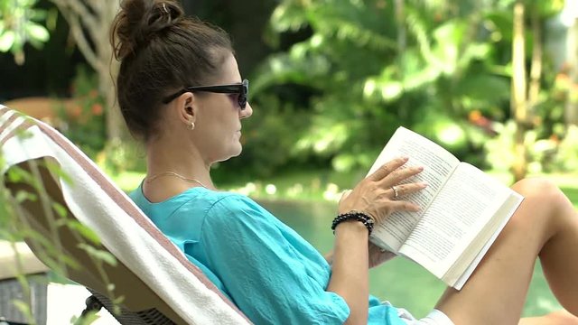 Woman yawning and looks bored while reading book next to the swimming pool, steadycam shot
