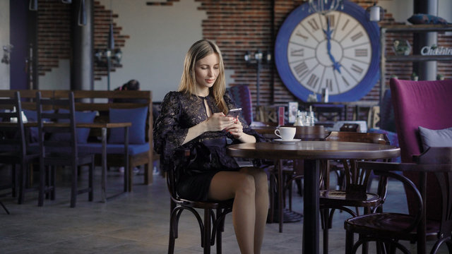 Businesswoman in black dress enjoying lunch break sitting in cafe with people in the background and using modern smartphone. Adorable lady relaxing in restaurant looking at digital device and smiling.
