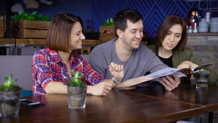 Three smiling people sitting in cafe and choosing food and beverages looking at the menu attentively. Friends spending their time together at the restaurant and discussing their dining.
