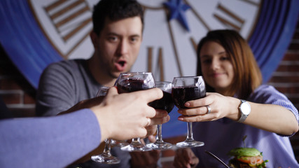 A group of cute young people in a modern restaurant at a table with snacks. Men and girls are baked with glasses of wine and shouting cheers. Friends celebrate a successful contract or birthday.
