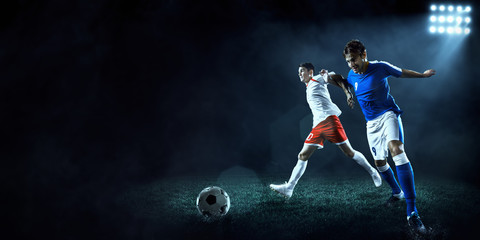 Plakat Soccer players performs an action play on a dark background. Soccer players fight for the ball. Players wears unbranded sport uniform.