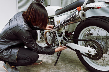 Fototapeta na wymiar Confident young man wearing black leather jacket repairing his vintage motorcycle while sitting on haunches at garage