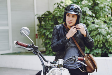 Pensive Asian biker wearing black leather jacket looking away while sitting on vintage motorcycle...