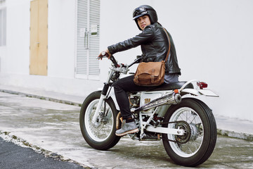 Obraz na płótnie Canvas Confident middle-aged biker wearing black leather jacket looking at camera while sitting on vintage motorcycle ready for searching adventures