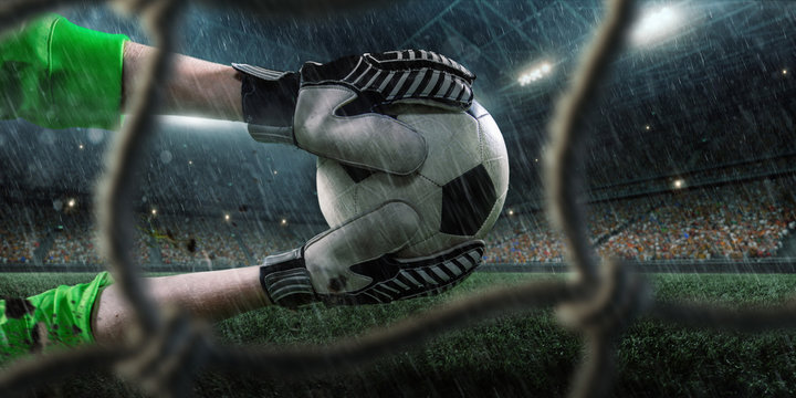 Soccer goalkeeper catches a ball on big professional rainy football arena. View through the football goal. Goalkeeper wears unbranded sport uniform.