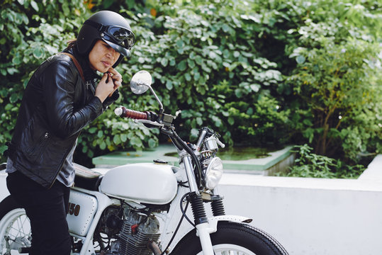 Brutal Middle-aged Biker Putting On Helmet And Looking At Rear View Mirror While Standing At Vintage Motorcycle Outdoors