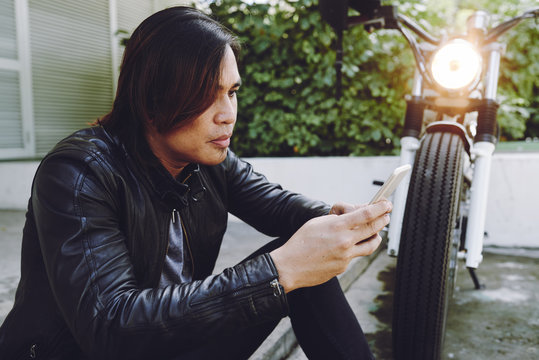 Concentrated Asian biker wearing black leather jacket studying electronic map on smartphone and sitting by side of road while taking break from motorcycle trip, profile view