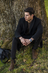Naklejka na ściany i meble Handsome young man leaning against tree in autumn, wearing black coat