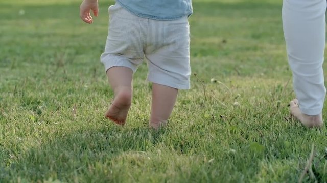Tilt up with rear view of toddler boy holding hand of unrecognizable mother and walking barefoot on green grass towards lake