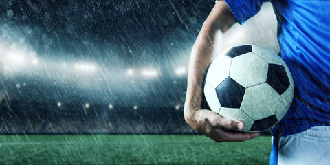 Soccer player holds a football ball on a professional rainy stadium. Player wears blue unbranded uniform.