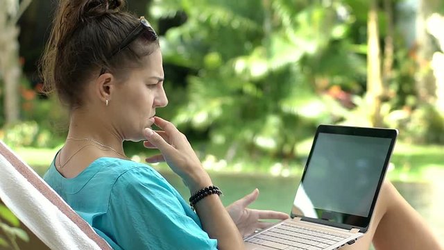 Woman looks irritated while using laptop next to the swimming pool, steadycam shot
