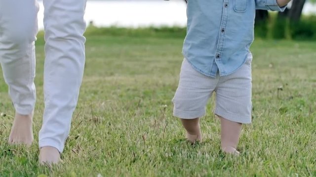 Tilt down of curious toddler boy holding hand of mother and walking barefoot on green grass on warm summer day