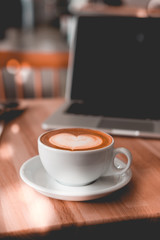 Mag of cappuccino with heart shaped latte art with laptop on background. Coffee shop wooden table. Freelancer, blogger working place concept