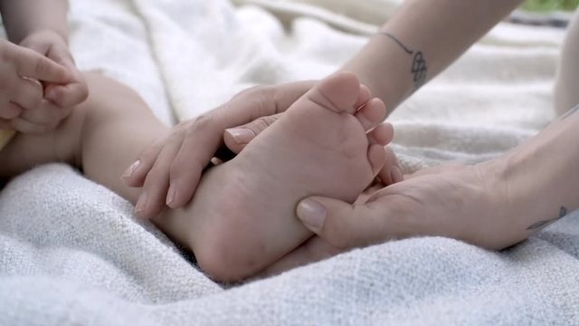 Close up of unrecognizable mother touching and massaging foot of baby boy sitting on towel outside