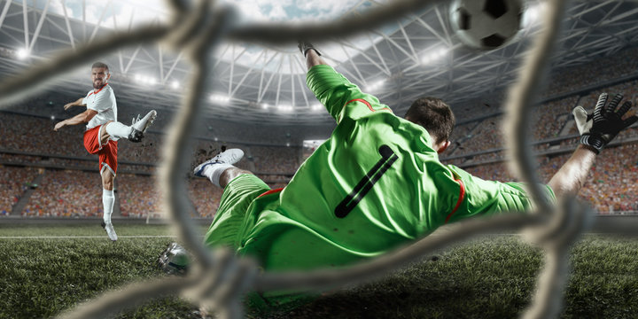 Soccer player scores the ball into the goal on professional stadium. The goalkeeper protects the football gate. Players wears unbranded sport uniform. View through the football gate.
