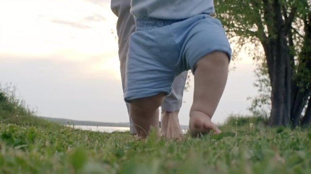 Low-section of laughing toddler and unrecognizable woman walking barefoot on green grass towards camera