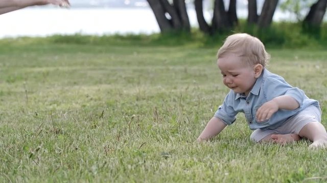 Tracking of adorable toddler boy with blond hair walking barefoot on green grass towards young mother, then falling down and crying