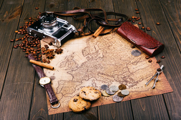 Old yellow map, glasses, coins, leather case, camera, watch, compasses, coffee beans, other spices and cookies lie on wooden floor