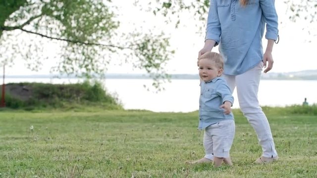 Tracking of unrecognizable mother holding hand of adorable toddler boy running barefoot on green grass in park with lake