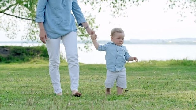 Lockdown of unrecognizable mother holding hand of cute little boy and walking on green grass in park on summer day; lake in background