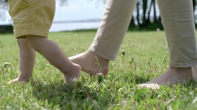 Tracking with low-section of legs of baby boy and unrecognizable woman walking on green grass