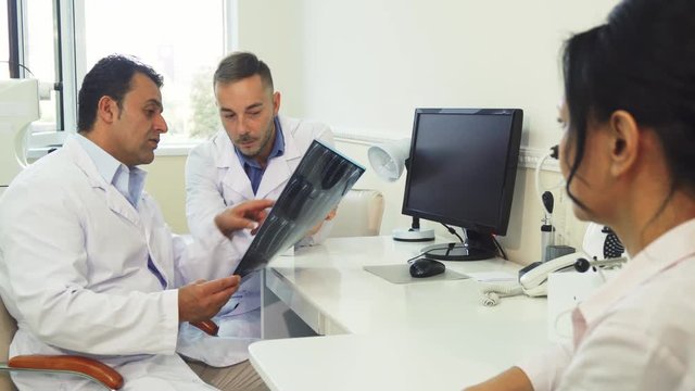 Two doctors and a patient are sitting in the office. One of them has X-Rays in their hands. They examine it and discuss it. The patient is waiting for results
