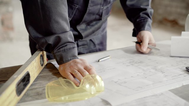 close up shot of at the man's hands, who makes notes in the construction drawing for the foreman and builders, the man is dressed in a protective uniform