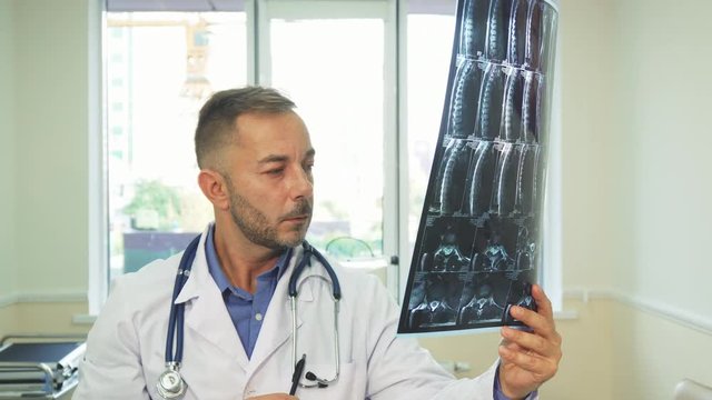 A man holding a picture of the X-Ray. He studies it very carefully. He has a stethoscope on his neck. He is in his office