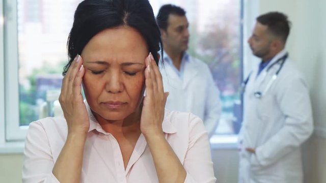 A woman is standing in the middle of the office. Suddenly she begins to feel pain in her temples. She puts her hands to her head. She looks very painful and unhappy