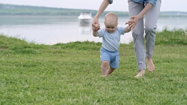 Unrecognizable woman holding hands of adorable baby boy walking on green grass towards camera