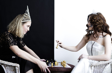 Two young women wearing queen dresses, diamond crown sitting playing chess. White and black background