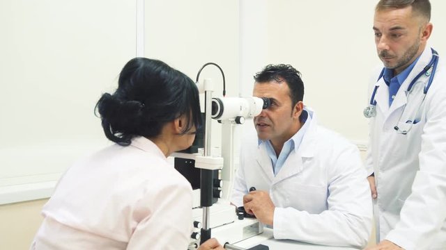 In the office are two doctors and a patient. One of the doctors checks the patients eyes with a special device. The second doctor closely observes these processes. They also have a nice conversation