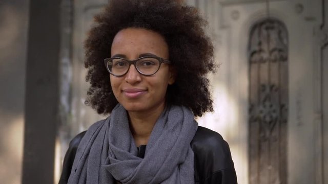 hister young metis woman with dark curly hair wearing glasses standing in front of old building in city street posing on camera with lovely smile slow motion