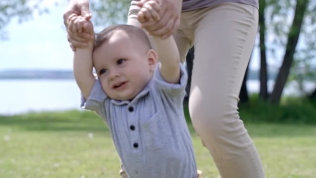 Unrecognizable mother holding hands of cute baby boy and helping him walk on green grass in park on summer day