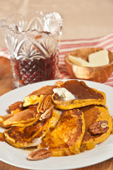 Stack of hot sweet potato pancakes, melting butter on a round, white plate, crystal pitcher of homemade maple syrup and a wood bowl of  butter, red and white stripped cloth