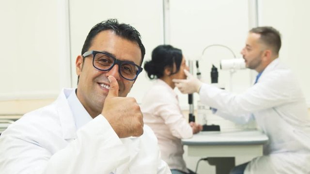 The doctor smiles and puts on his glasses. He shows his thumb up. In the background is another doctor. He makes the diagnosis of the patients eyes