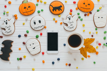 A preparation for Halloween: Cup of coffee and smartphone on the white wooden background with candy and gingerbread.